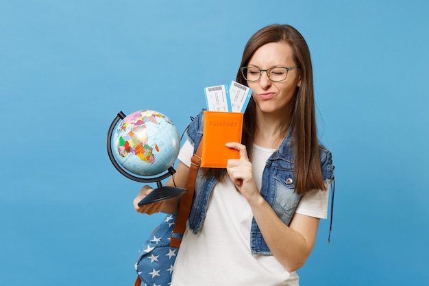 Young funny woman student in glasses with backpack holding world glove, passport, boarding pass tickets isolated on blue background. Education in university college abroad. Air travel flight concept.