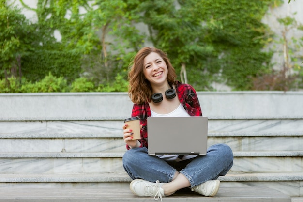노트북을 사용하고 야외에서 커피를 마시는 재미있는 젊은 여성 학생. 원격 근무 또는 교육 개념