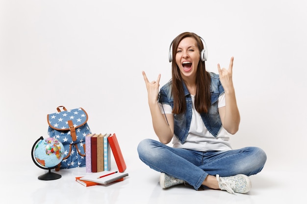 Young funny woman student with headphones listening music showing rock-n-roll sign screaming near globe backpack school books isolated on white wall