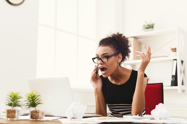 Young furious african-american woman talking on phone at modern office copy space. Business consulting.