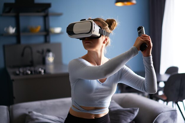 Young futuristic blonde girl wearing virtual reality headset holding controller playing a video game