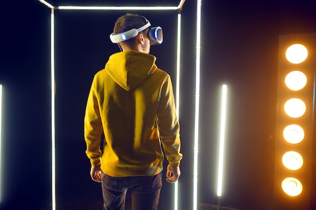 Молодой геймер играет в игру, используя шлем виртуальной реальности и геймпад в светящемся кубе. Темный интерьер игрового клуба, технология VR с 3D-зрением