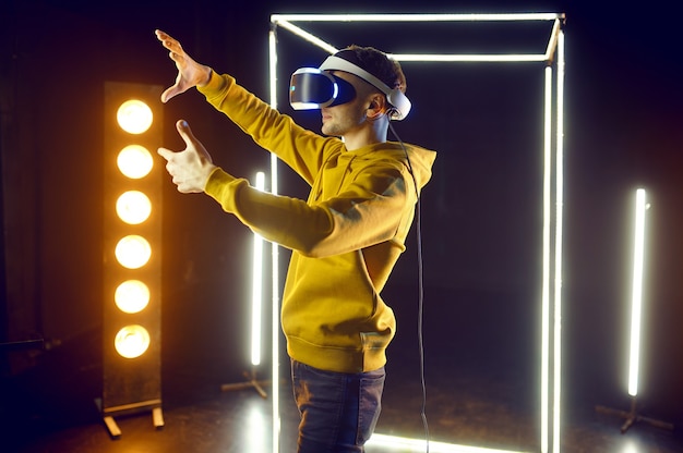 Фото Молодой геймер играет в игру, используя гарнитуру виртуальной реальности и геймпад в светящемся кубе, вид спереди. темный интерьер игрового клуба, технология vr с 3d-зрением