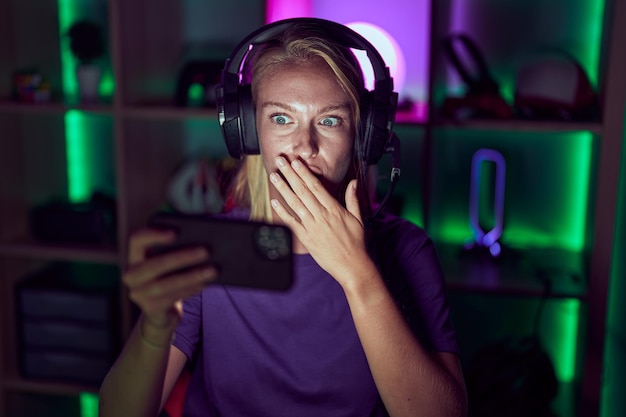 Young gamer woman playing video games with smartphone covering mouth with hand, shocked and afraid for mistake. surprised expression