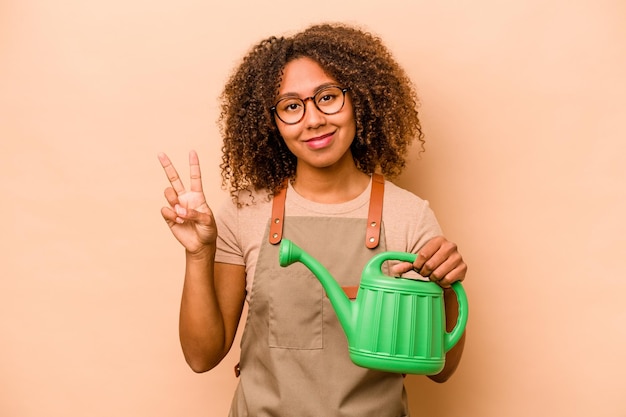 Young gardener African American woman holding irrigation isolated on beige background showing number two with fingers
