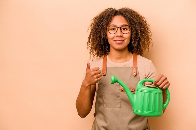 Young gardener African American woman holding irrigation isolated on beige background smiling and raising thumb up