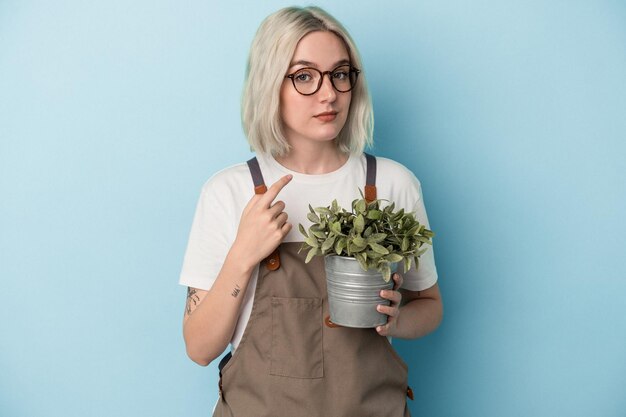 誘うようにあなたに指で指している青い背景に分離された植物を保持している若い庭師白人女性が近づく