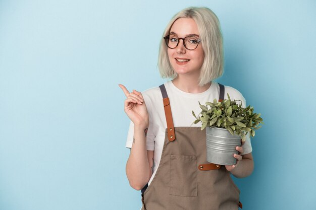 Young gardener caucasian woman holding a plant isolated on blue background smiling and pointing aside, showing something at blank space.