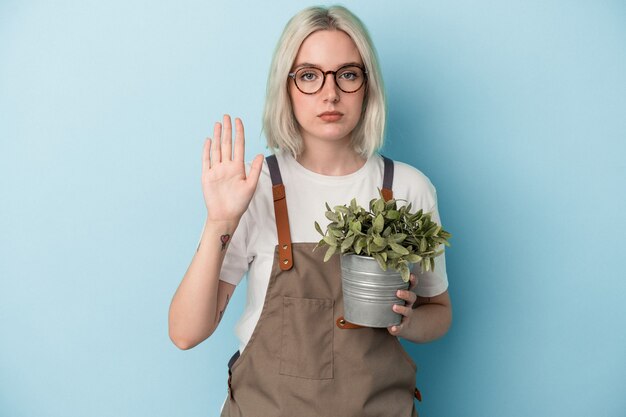 一時停止の標識を示す伸ばした手で立っている青い背景で隔離の植物を保持している若い庭師の白人女性は、あなたを防ぎます。