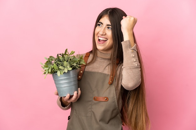 Young gardener girl holding a plant isolated on pink background celebrating a victory