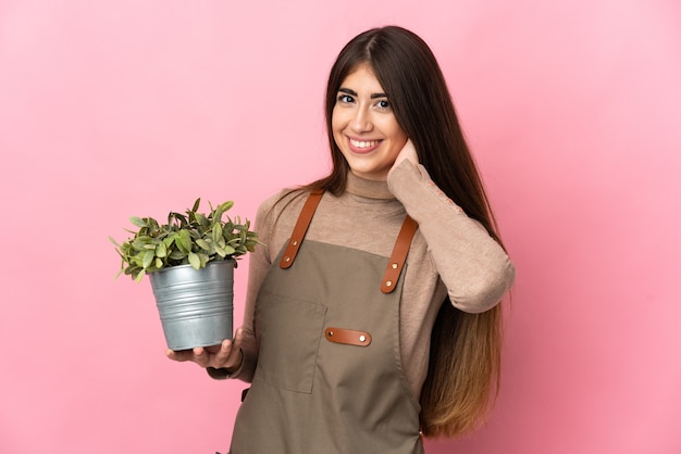 Young gardener girl holding a plant isolated on pink background laughing