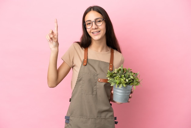 素晴らしいアイデアを指しているピンクの背景に分離された植物を保持している若い庭師の女の子