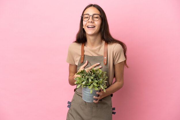 たくさん笑ってピンクの背景に分離された植物を保持している若い庭師の女の子