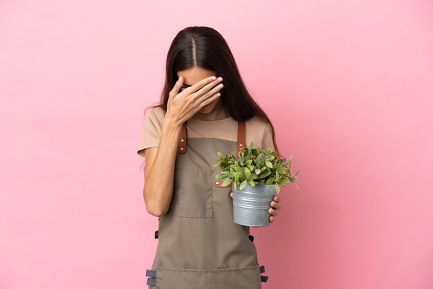 疲れて病気の表情でピンクの背景に分離された植物を保持している若い庭師の女の子