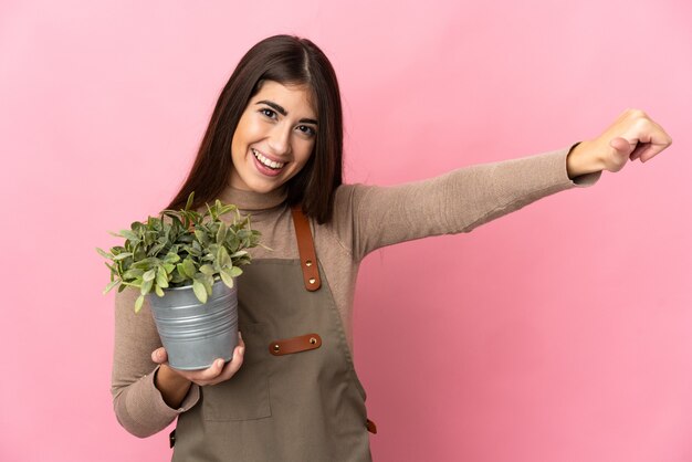 Young gardener girl holding a plant isolated on pink wall giving a thumbs up gesture