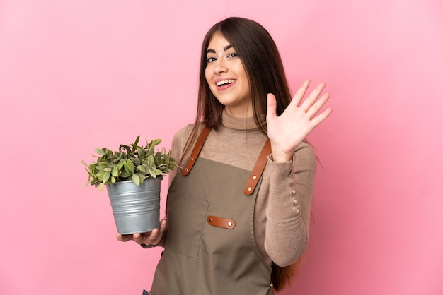 Giovane ragazza del giardiniere che tiene una pianta isolata che saluta con la mano con l'espressione felice