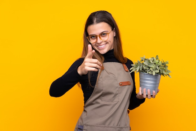 Young gardener girl holding a plant points finger at you