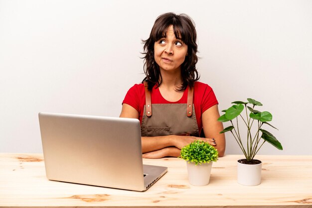 Young gardener hispanic woman sitting on a table isolated on white background confused feels doubtful and unsure