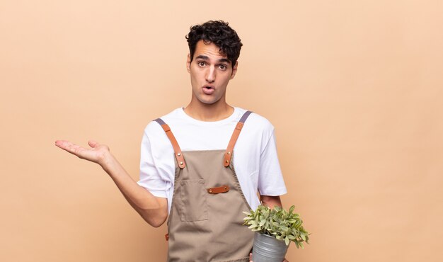 Young gardener man looking surprised and shocked, with jaw dropped holding an object with an open hand on the side