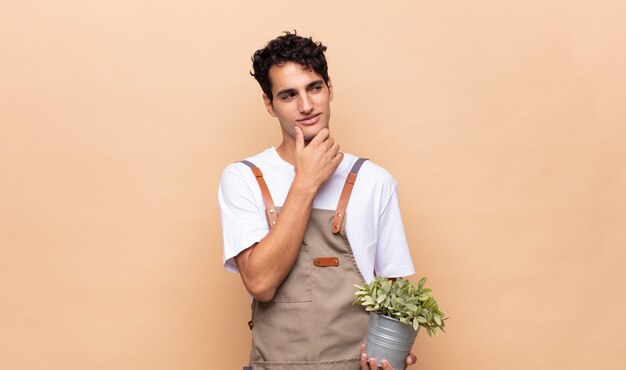 Young gardener man smiling with a happy, confident expression with hand on chin, wondering and looking to the side