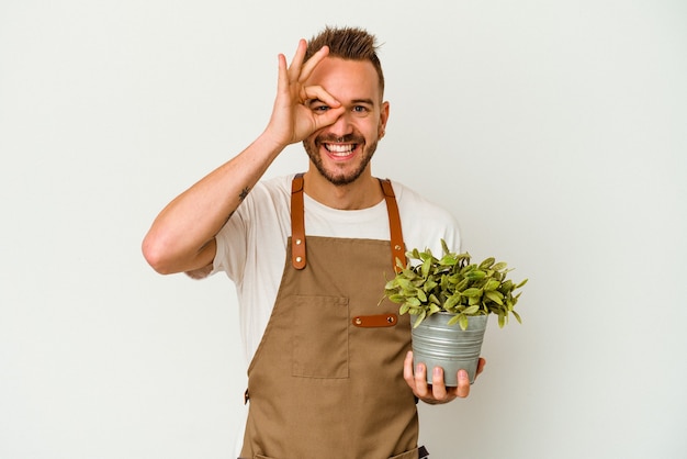Молодой садовник татуированный кавказский мужчина, держащий растение, изолированное на белом фоне, взволнован, держа на глазах хорошо жест.