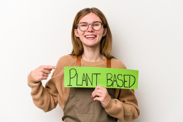 Young gardener woman holding a plan based placard isolated on white background smiling and pointing aside showing something at blank space