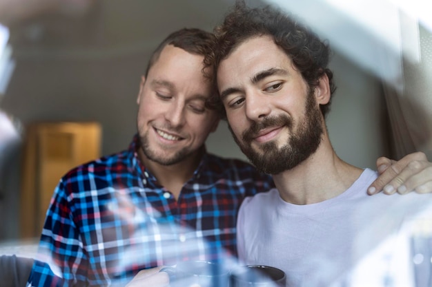 Giovane coppia gay innamorata che guarda fuori dalla finestra due giovani uomini androgini che sorridono insieme e bevono caffè