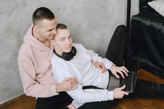 Young gay couple sitting on floor using laptop, using headphones listen to music together, hugging or embracing.