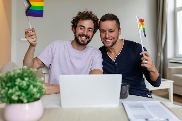 LGBTQ 깃발을 들고 화상 통화로 친구들과 인사하면서 유쾌하게 웃고 있는 젊은 게이 커플