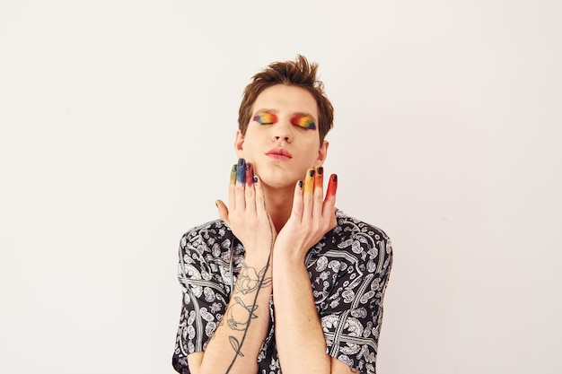 Young gay man is standing in the studio and posing for a camera Multicolored make up on face fingers