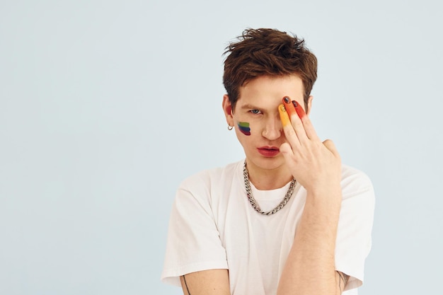 Young gay man is standing in the studio and posing for a camera Painted by multi colored makeup