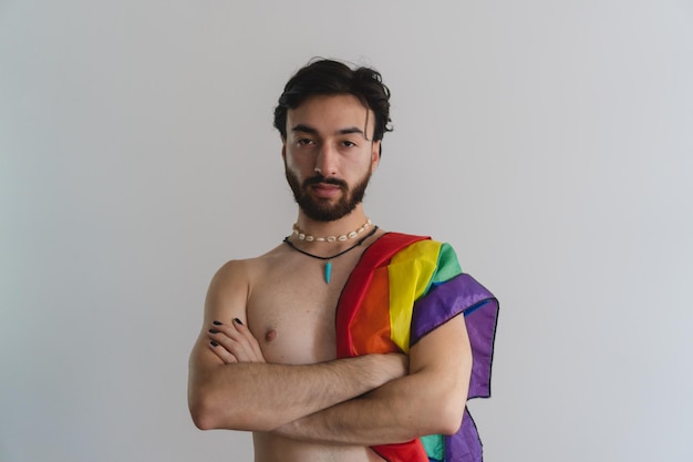 Young gay queer latin man with arms crossed looking at camera with lgtb flag over his shoulder