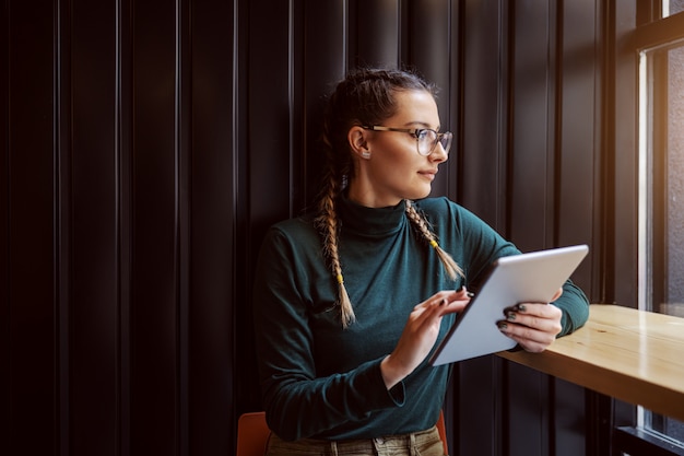 Giovane ragazza geniale che si siede nella caffetteria accanto alla finestra, utilizzando tablet per navigare in internet e guardando attraverso la finestra.