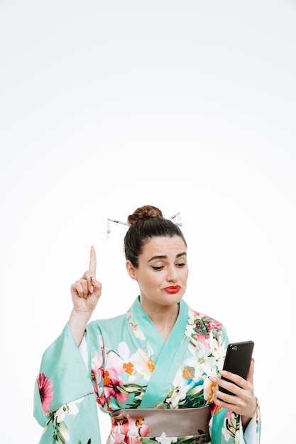 young geisha woman in traditional japanese kimono holding smartphone looking at it with skeptic expression pointing with index finger up have a idea standing over white wall