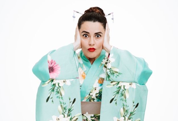 young geisha woman in traditional japanese kimono looking at front being stressed and shocked covering ears with hands standing over white wall