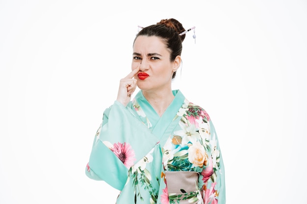 young geisha woman in traditional japanese kimono looking at front confused touching her nose standing over white wall