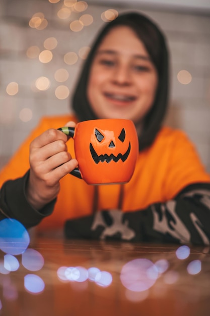 Young generations Z teen boy in orange clothes while all hallows eve photo session at home Stylish teenager with jack lantern mug and sweets celebrates halloween enjoy chocolate candy