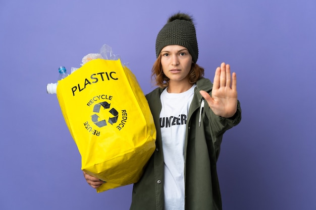 Foto giovane ragazza georgiana che tiene un sacchetto pieno di bottiglie di plastica per riciclare facendo il gesto di arresto