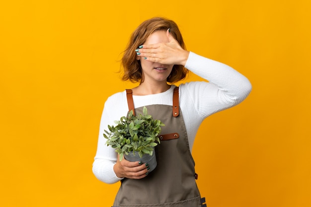 手で目を覆っている黄色の壁に隔離された植物を保持している若いグルジアの女性。何かを見たくない