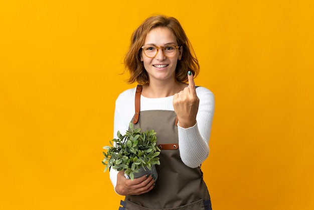 来るジェスチャーをしている黄色の壁に隔離された植物を保持している若いジョージ王朝の女性