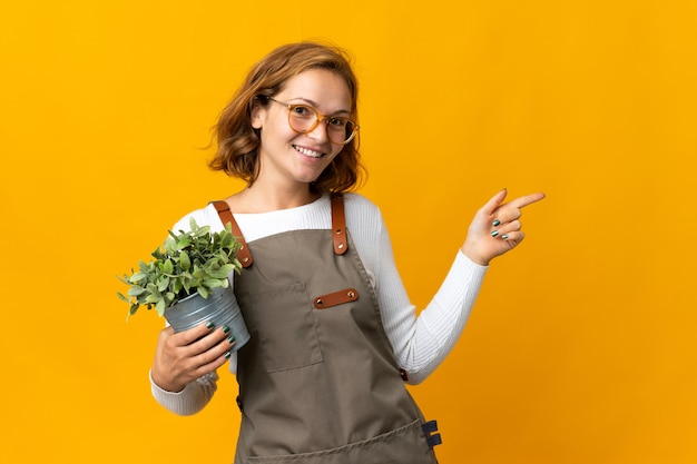 横に指を指している黄色の壁に隔離された植物を保持している若いジョージ王朝様式の女性