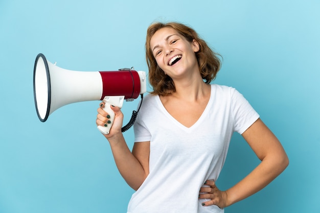 Young Georgian woman isolated on blue wall holding a megaphone and smiling