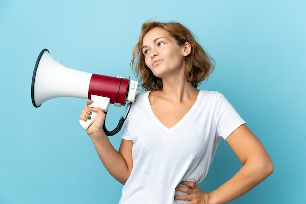 Young Georgian woman isolated on blue wall holding a megaphone and thinking