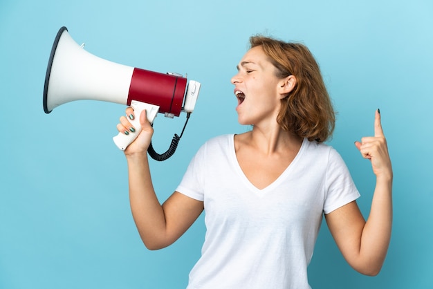 Young Georgian woman isolated on blue wall shouting through a megaphone to announce something in lateral position