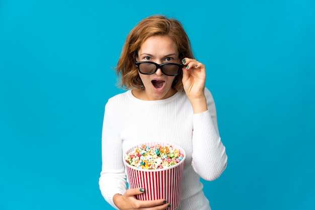 Young Georgian woman isolated on blue wall surprised with 3d glasses and holding a big bucket of popcorns