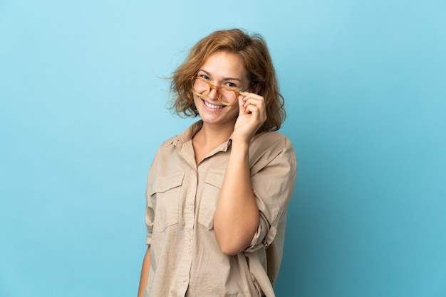 Young Georgian woman isolated on blue with glasses and happy