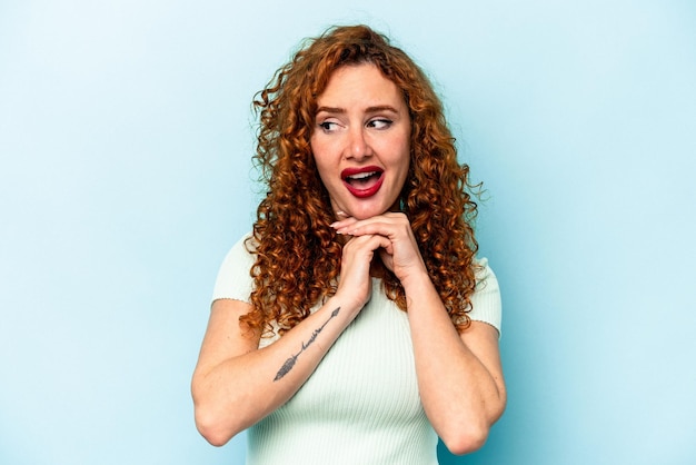 Young ginger caucasian woman isolated on blue background praying for luck amazed and opening mouth looking to front