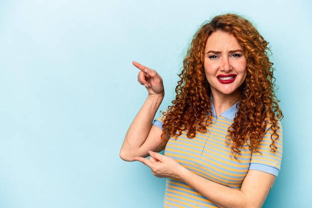 Young ginger caucasian woman isolated on blue background shocked pointing with index fingers to a copy space