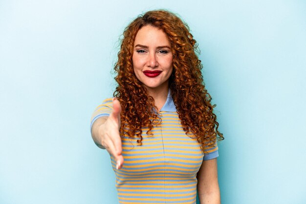 Young ginger caucasian woman isolated on blue background stretching hand at camera in greeting gesture