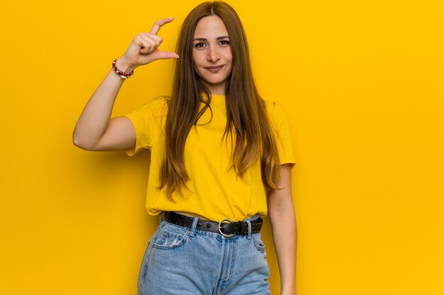 Young ginger redhead woman holding something little with forefingers, smiling and confident